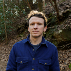 Man in blue coat standing in the woods looking into the camera.